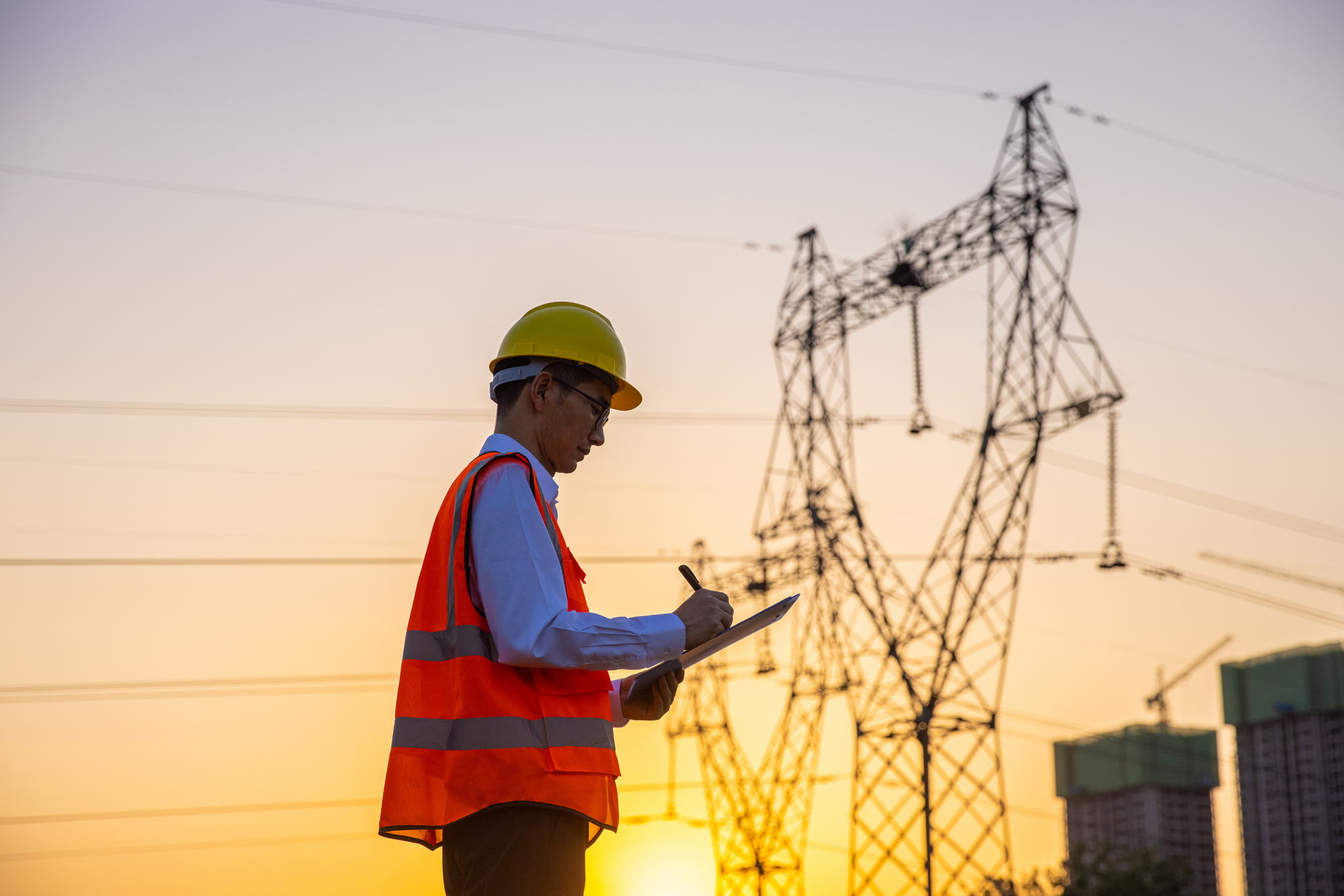 An engineer wearing a yellow hard hat and orange safety vest writes on a clipboard while standing near high-voltage power lines at sunset, with construction cranes visible in the background.