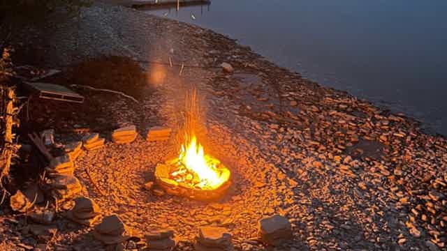 Campfire on rocky shore, flames rising from a ring of stones, nearby dark water.