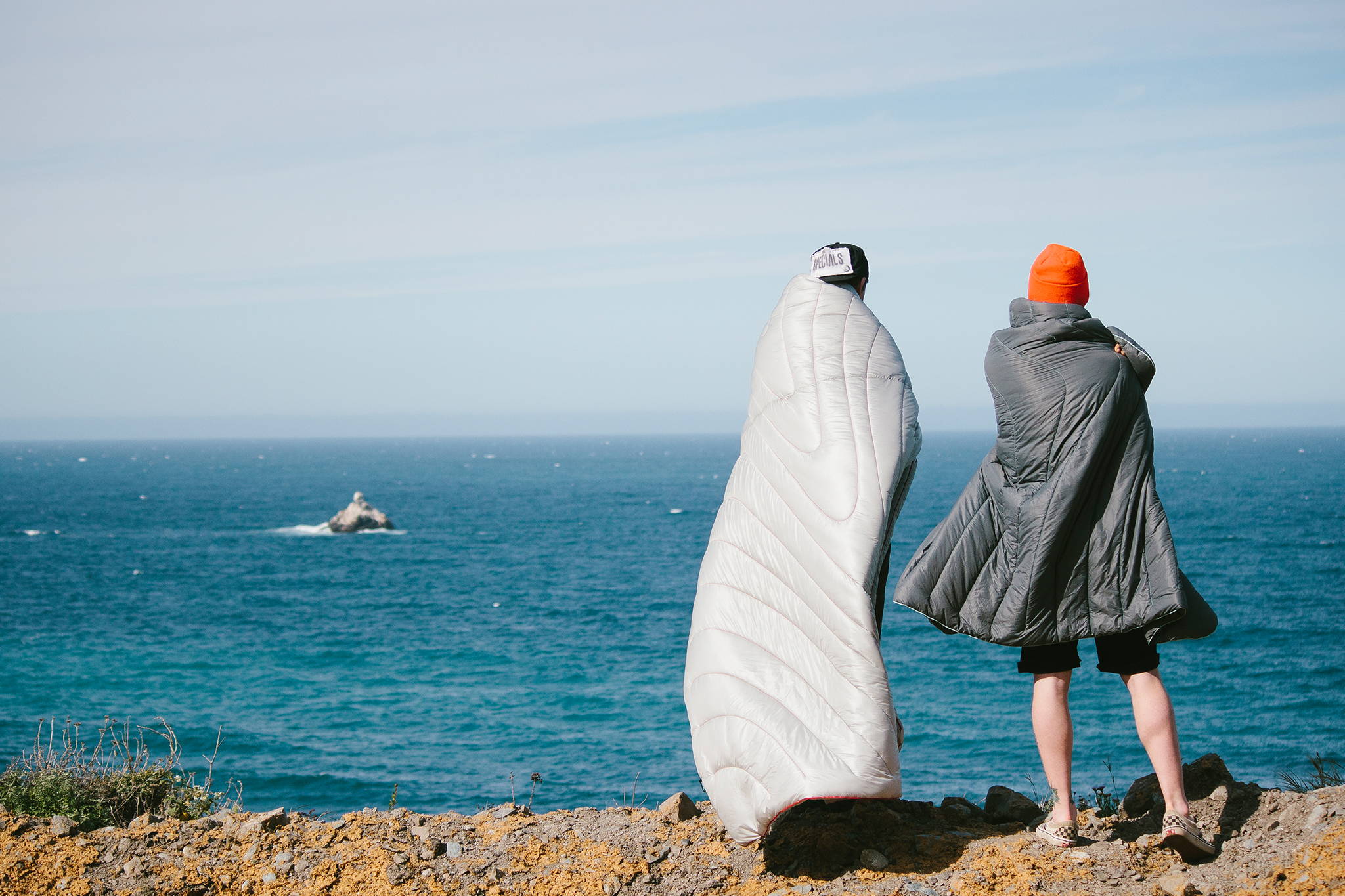 Two people standing looking at the ocean wearing Rumpl outdoor blankets