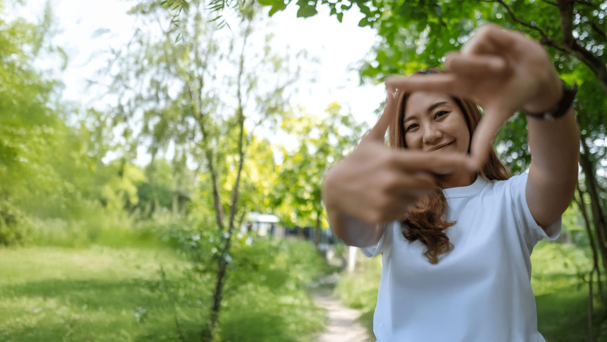 A smiling woman in a white shirt stands outside on a sunny day, holding her hands up to form a rectangular frame. She is surrounded by green trees and grass, with a blurred path in the background.