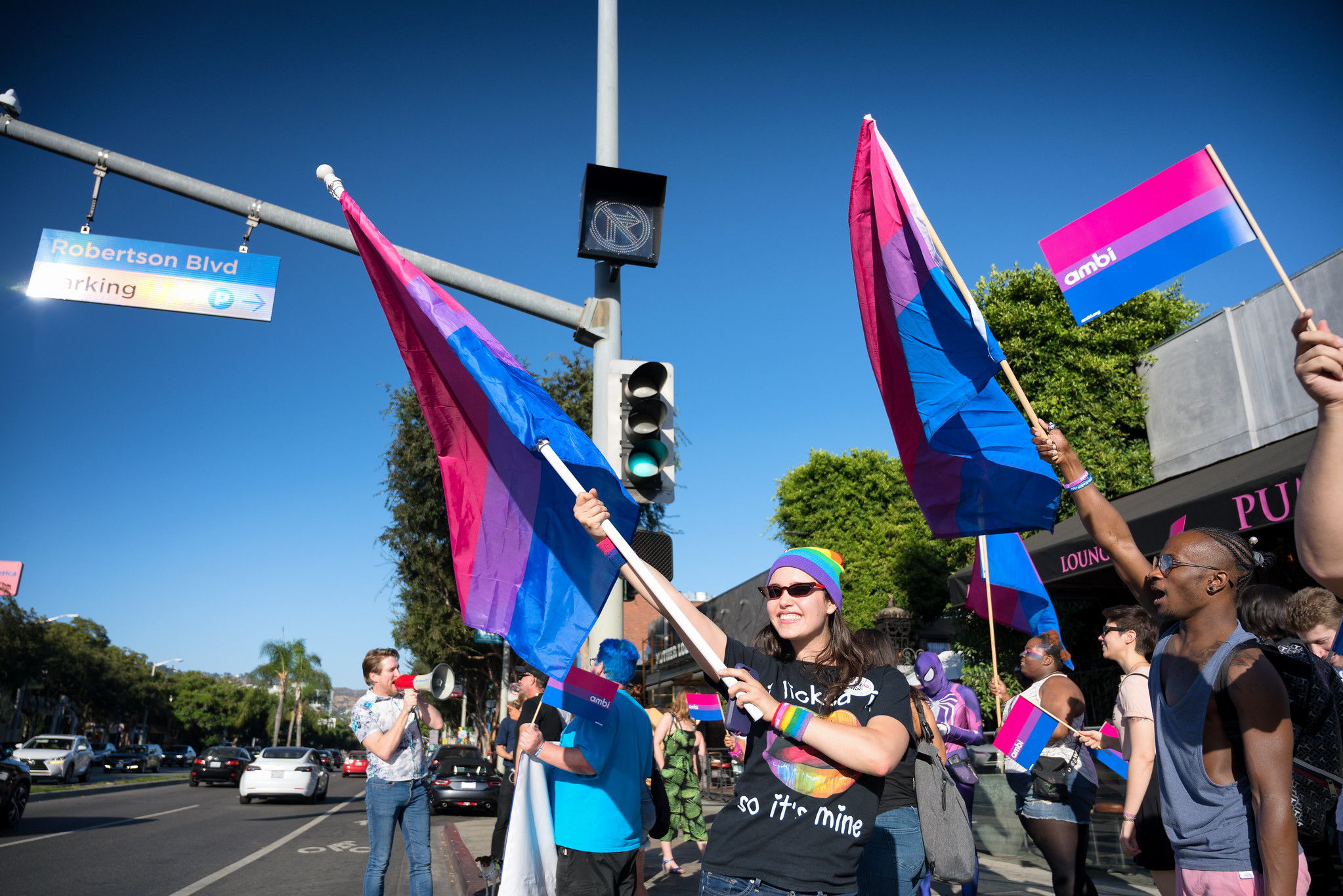 People on the sidewalk smiling wearing pride shirts and holding purple bi flags.