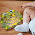Baby sitting next to a colorful frog-shaped magnetic maze with a cover and a magnetic pen.