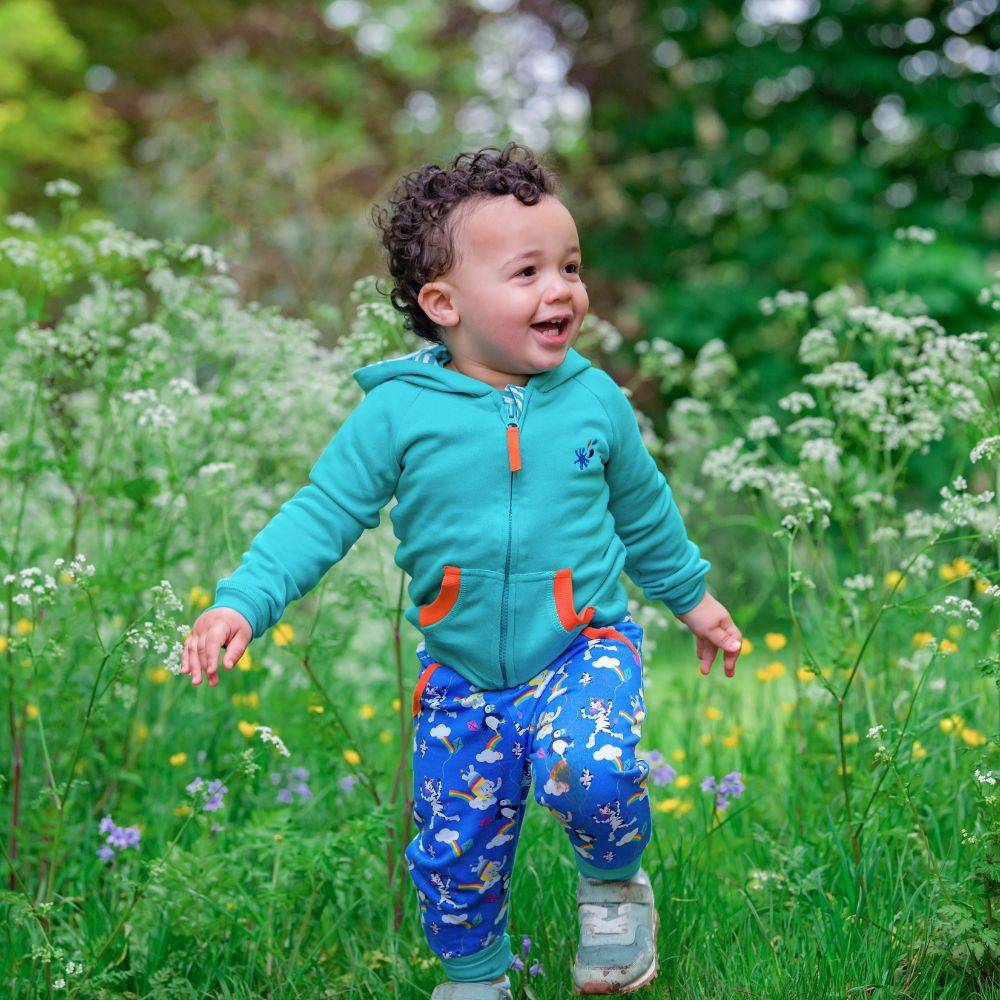 Image of a smiling toddler surrounded by long grass and wild flowers, wearing a Ducky Zebra turquoise hoody and brightly coloured Ducky Zebra trousers