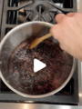 A hand stirs a dark mixture with a wooden spoon in a saucepan on a stove.
