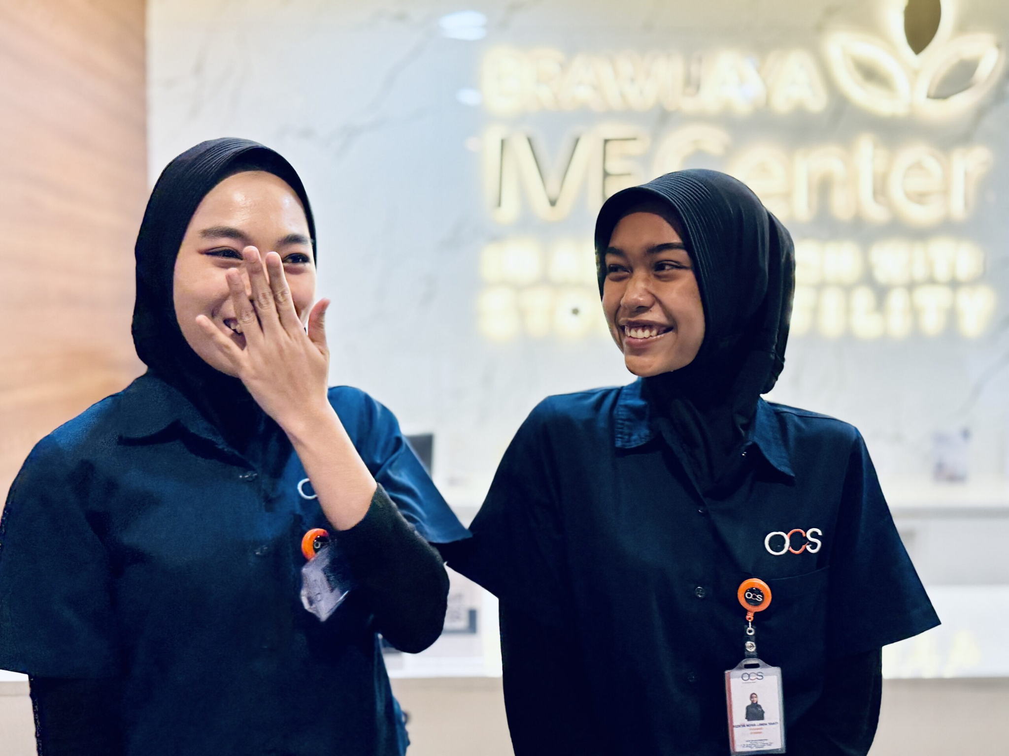 Two women wearing black hijabs and navy blue uniforms smile and stand together inside a clinic or medical center. One woman covers her mouth while laughing. The blurred background shows a sign that reads IVF Center.