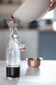 A hand pours a dark liquid from a white container through a funnel into a glass bottle on a kitchen counter.