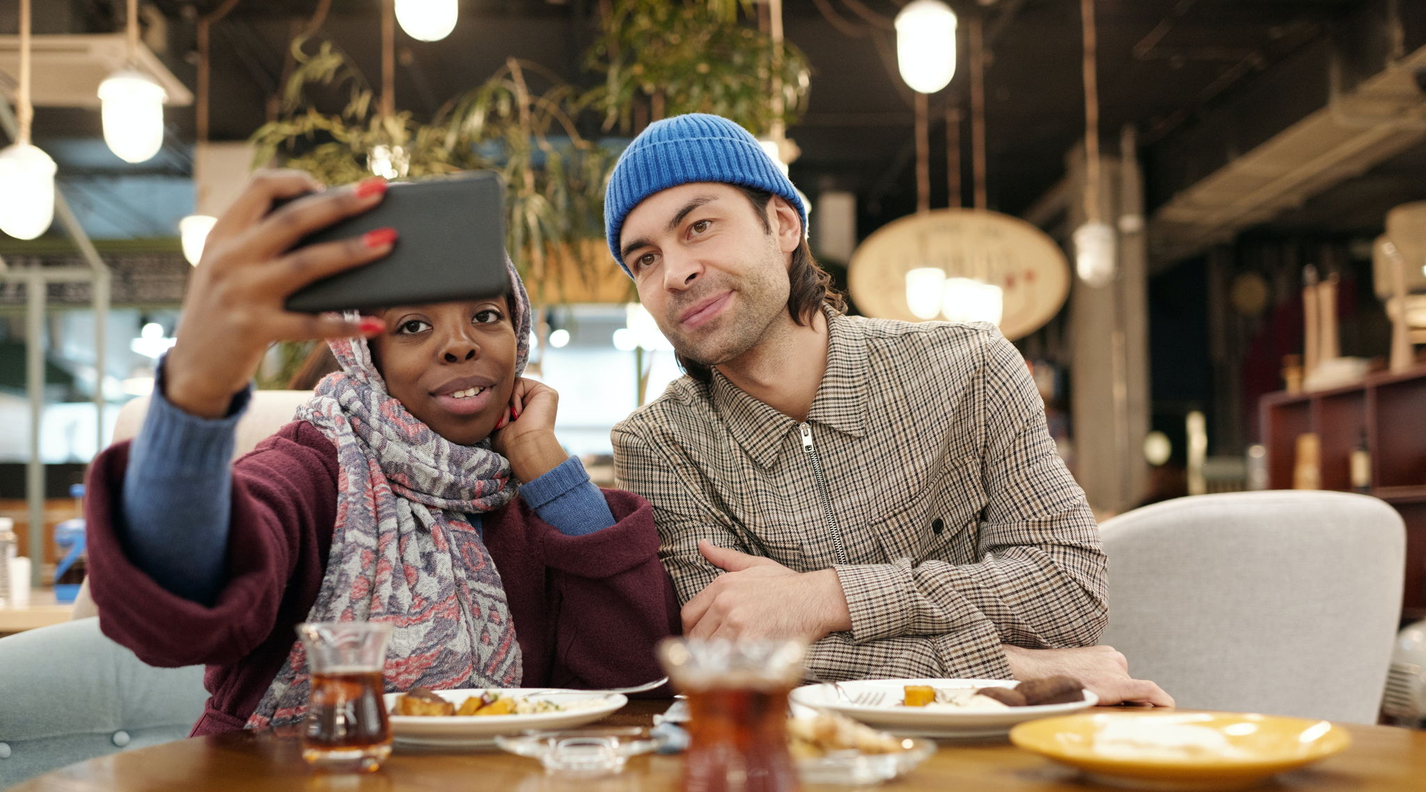 A woman wearing a head scarf and a hipster guy take a selfie together while they eat breakfast at a diner.