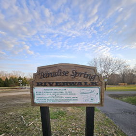 Wooden sign "Paradise Spring Riverwalk" in a serene natural landscape, suggesting a peaceful walking trail near water.