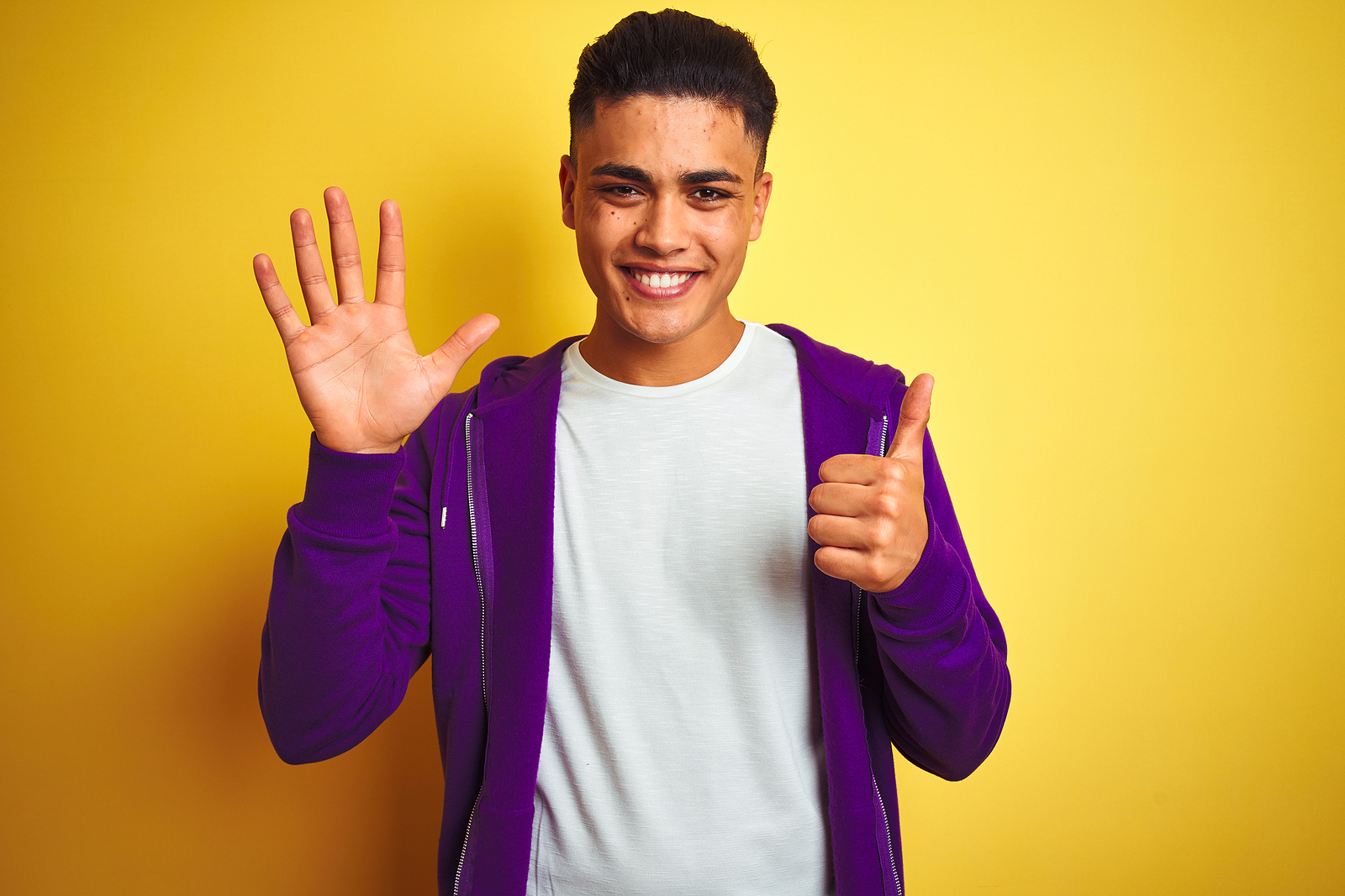A young latino man smiles while holding 6 fingers up against a plain background. He has on a purple sweater.