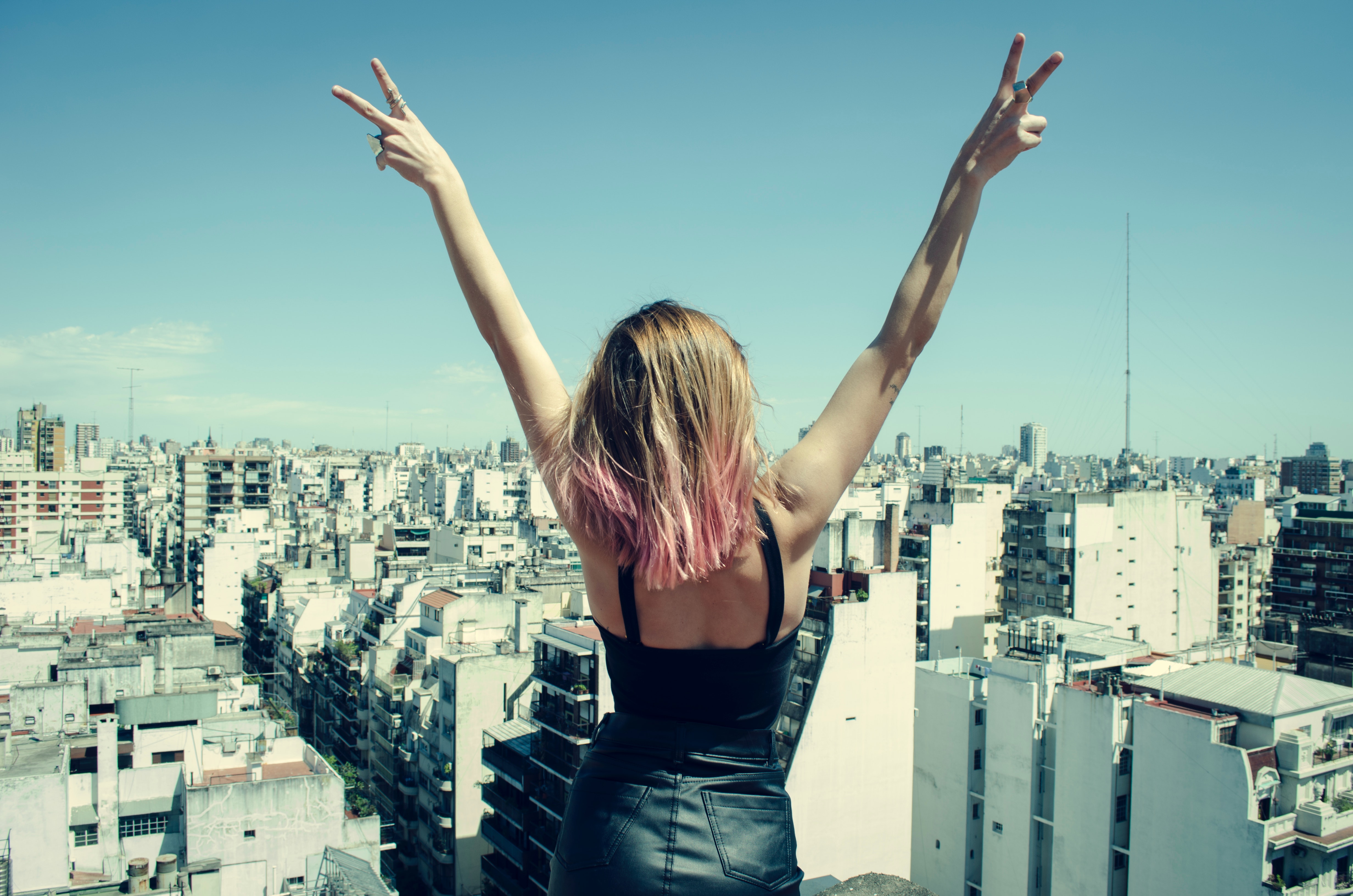 A confident woman wearing a leather dress holds her hands in a peace sign at the top of a building.