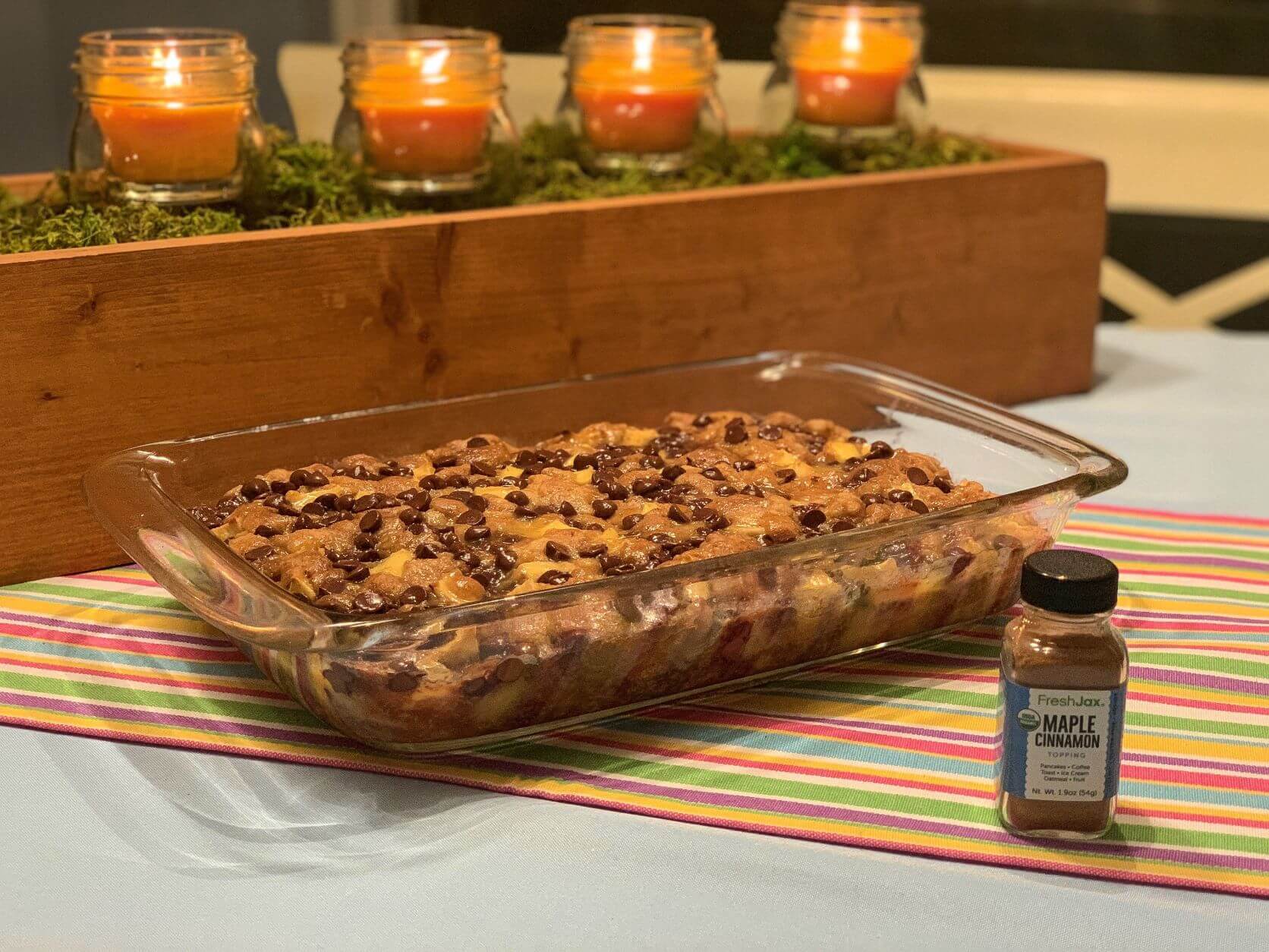 Chocolate chip apple cake in glass dish with a sampler bottle of FreshJax maple cinnamon topping. Orange candles in background. 