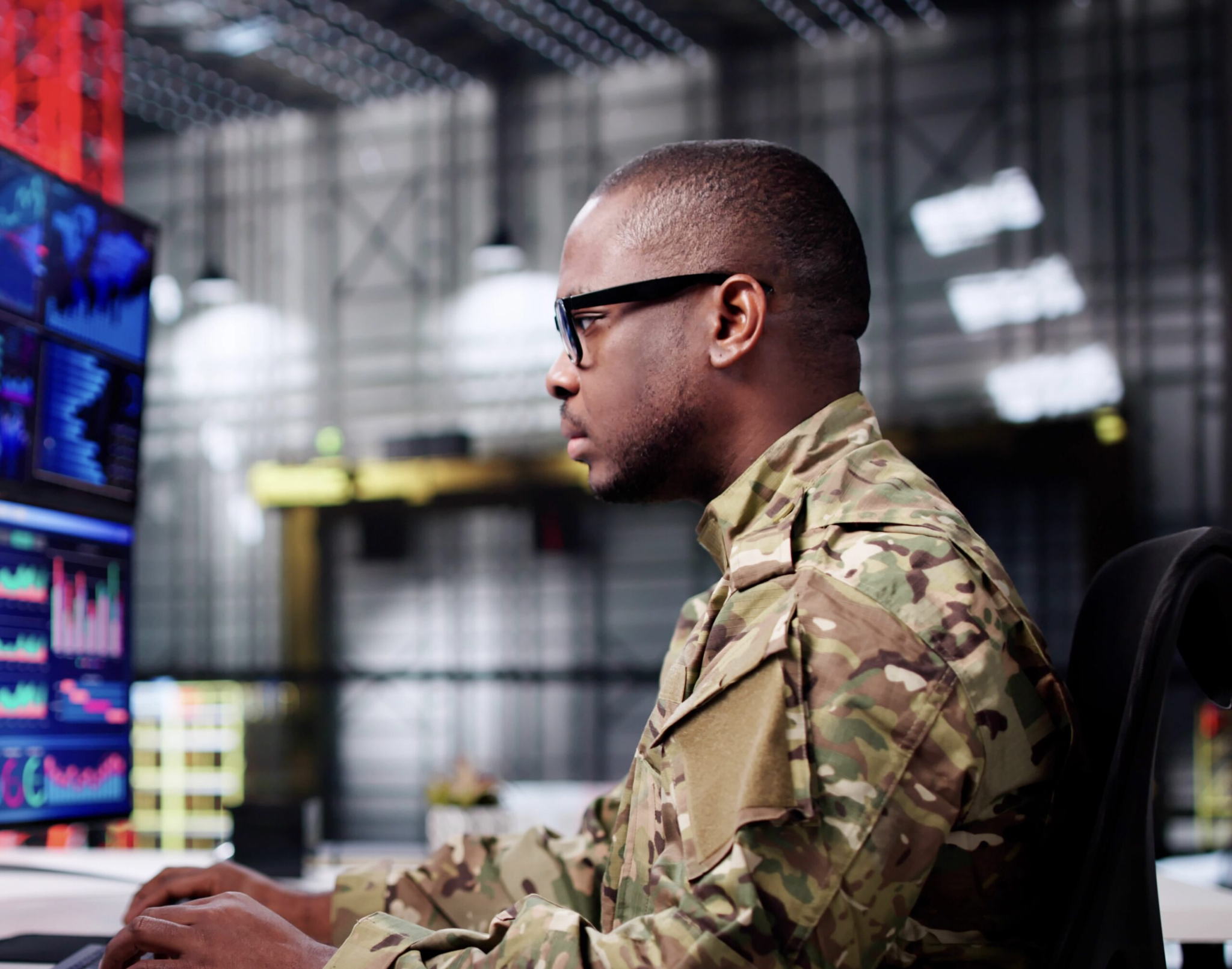 A man in military camouflage uniform and glasses works at a computer in a high-tech control room with multiple monitors displaying data and graphs.