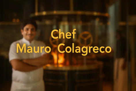 Chef Mauro Colagreco smiles confidently with arms crossed, wearing a white chef's jacket in front of a large, warm oven in a restaurant kitchen.