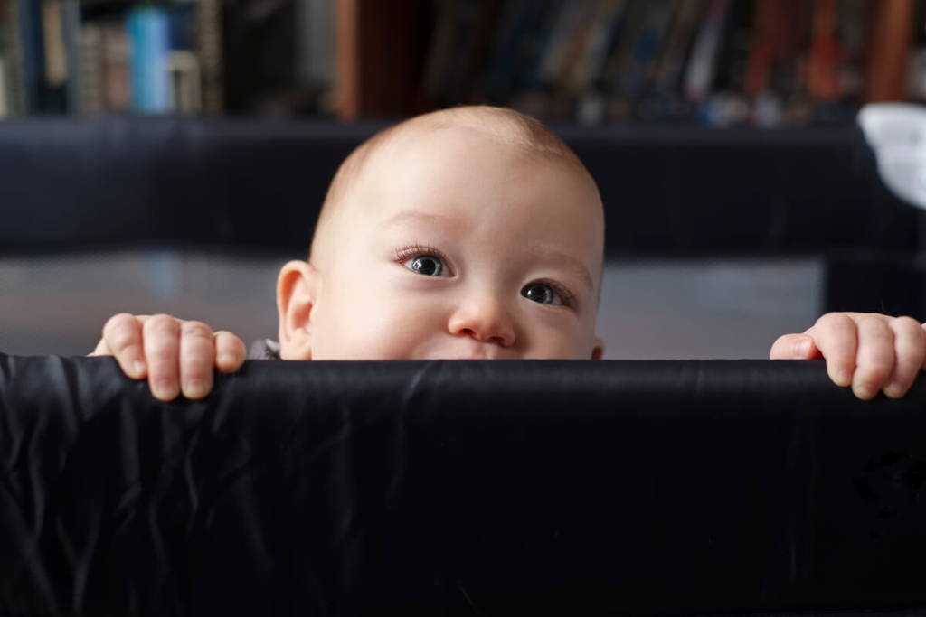 Un bébé curieux regarde par-dessus le bord de son parc, s'accrochant de ses deux mains et fixant l'horizon avec de grands yeux.