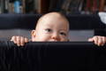 Curious baby peeking over the edge of a playpen, holding on with both hands and gazing forward with wide eyes.