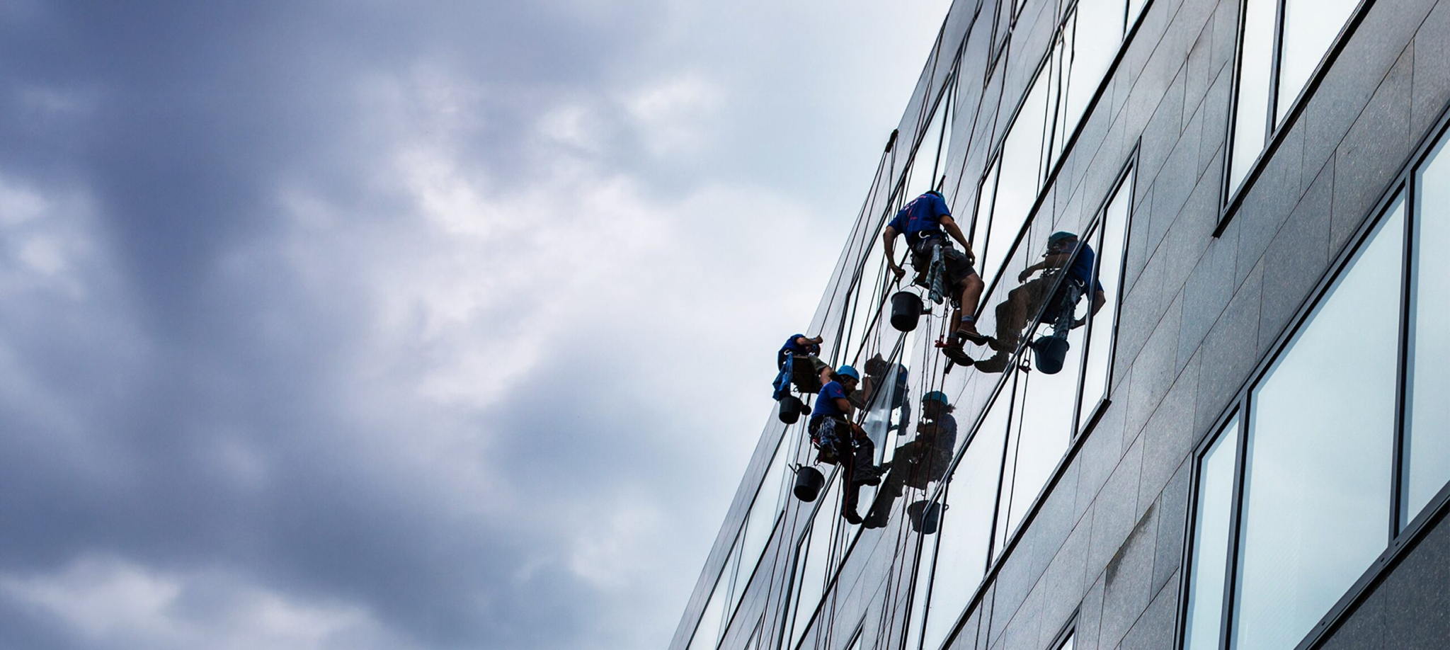 Four window cleaners wearing safety gear are suspended by ropes, cleaning the glass windows of a tall building under a cloudy sky.