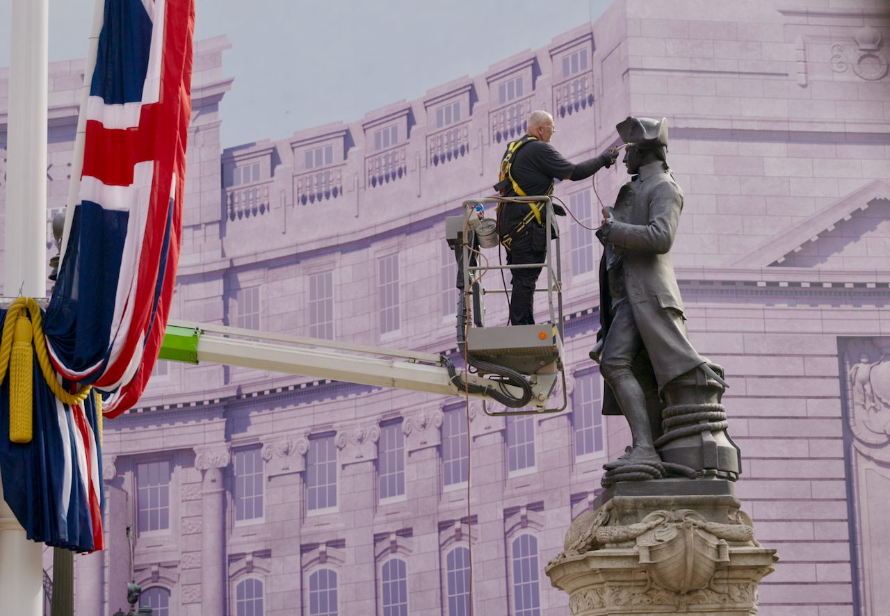 A worker on a mechanical lift cleans or repairs a large statue while wearing safety gear. A draped British flag is visible to the left, and an ornate stone building stands in the background.