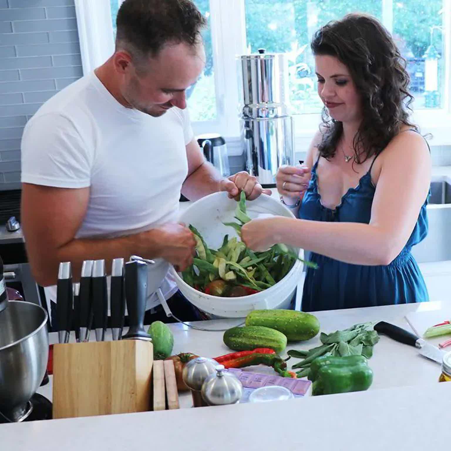 Two people chop fresh vegetables in a bright kitchen, mixing greens in a large bowl with various produce on the counter.