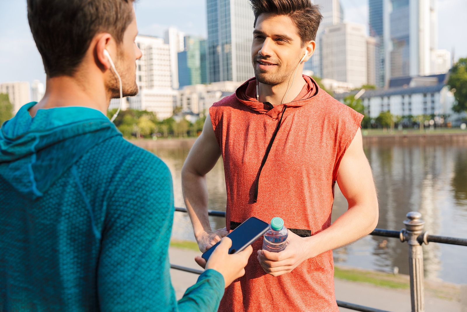 Photo of 2 young white guys with workout clothes both talking to each other near a lake in the city.