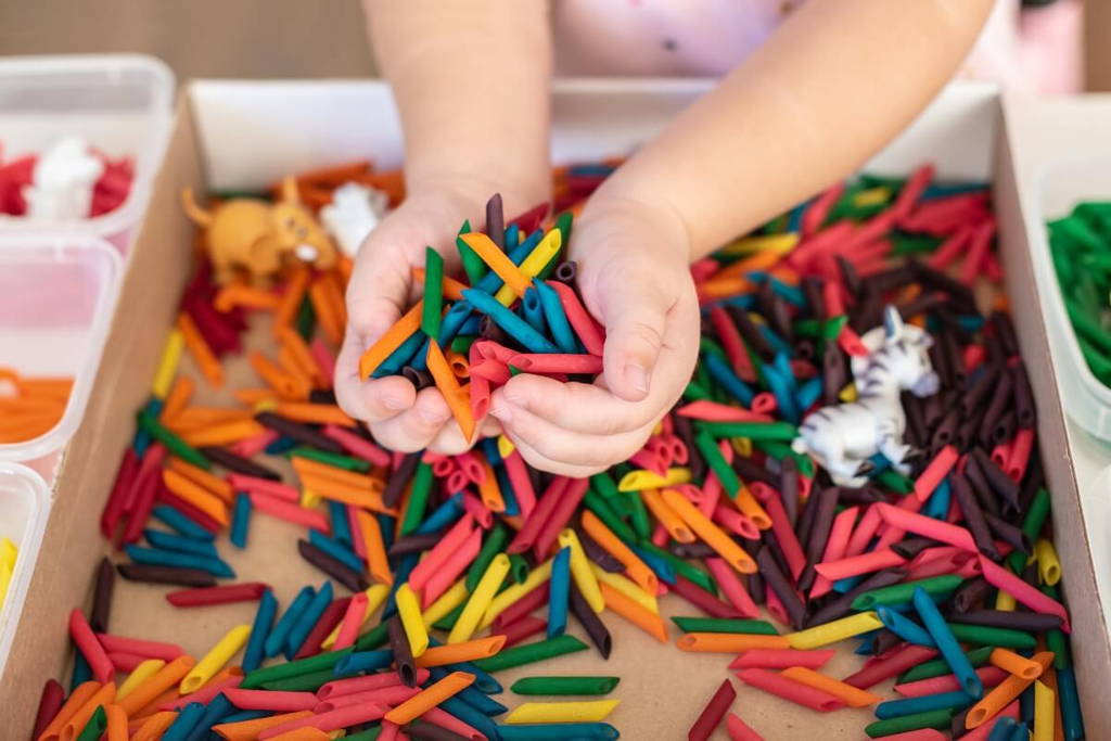 Child's hands grabbing colorful pasta from the sensory bin.