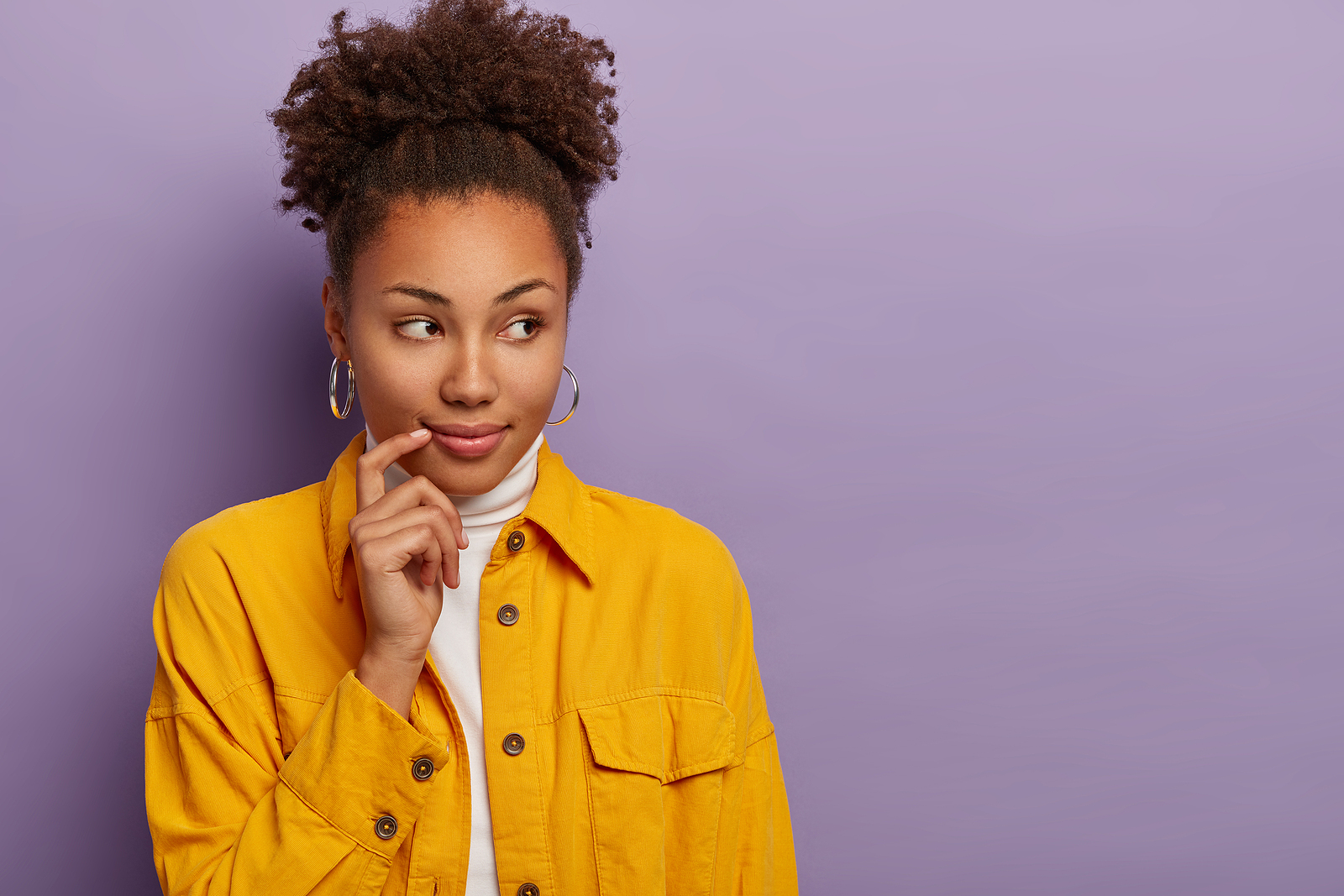 A woman with a dark complexion smiles curiously and looks to the side holding a finger up to her cheek in front of a purple background.