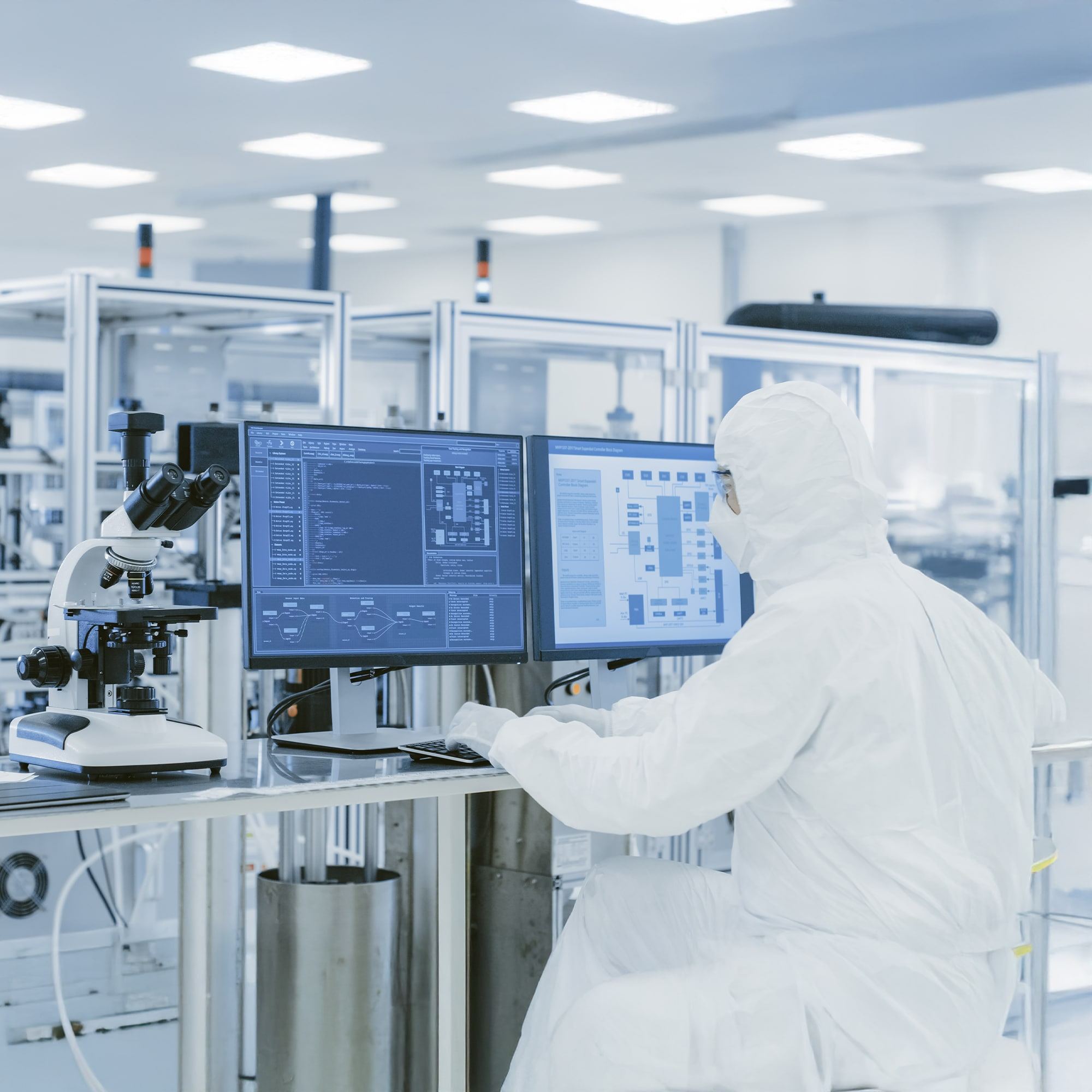 A scientist in full protective gear works at a dual-monitor computer station in a cleanroom laboratory, with a microscope nearby and advanced equipment in the background.