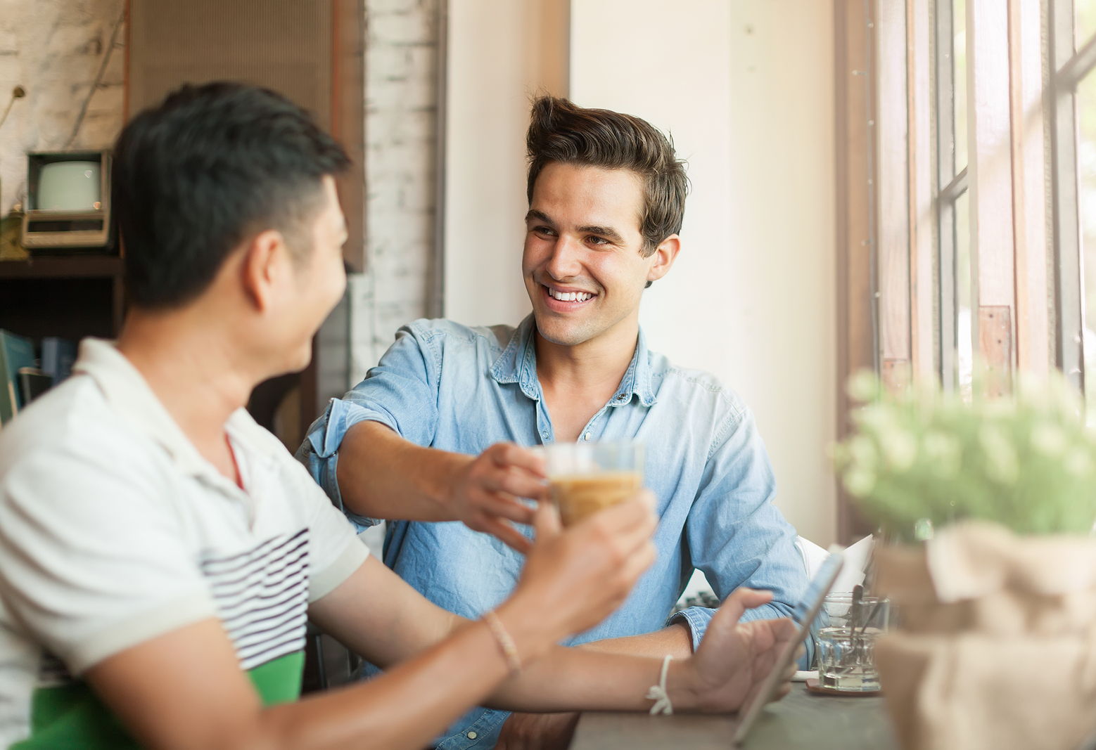 Two friends talking and about to cheer their coffee mugs together while they smile.