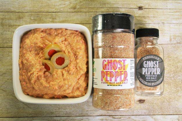 A bowl of spicy olive hummus next to a large and small bottle of FreshJax Organic Ghost Pepper Fiery Hot Sea Salt.