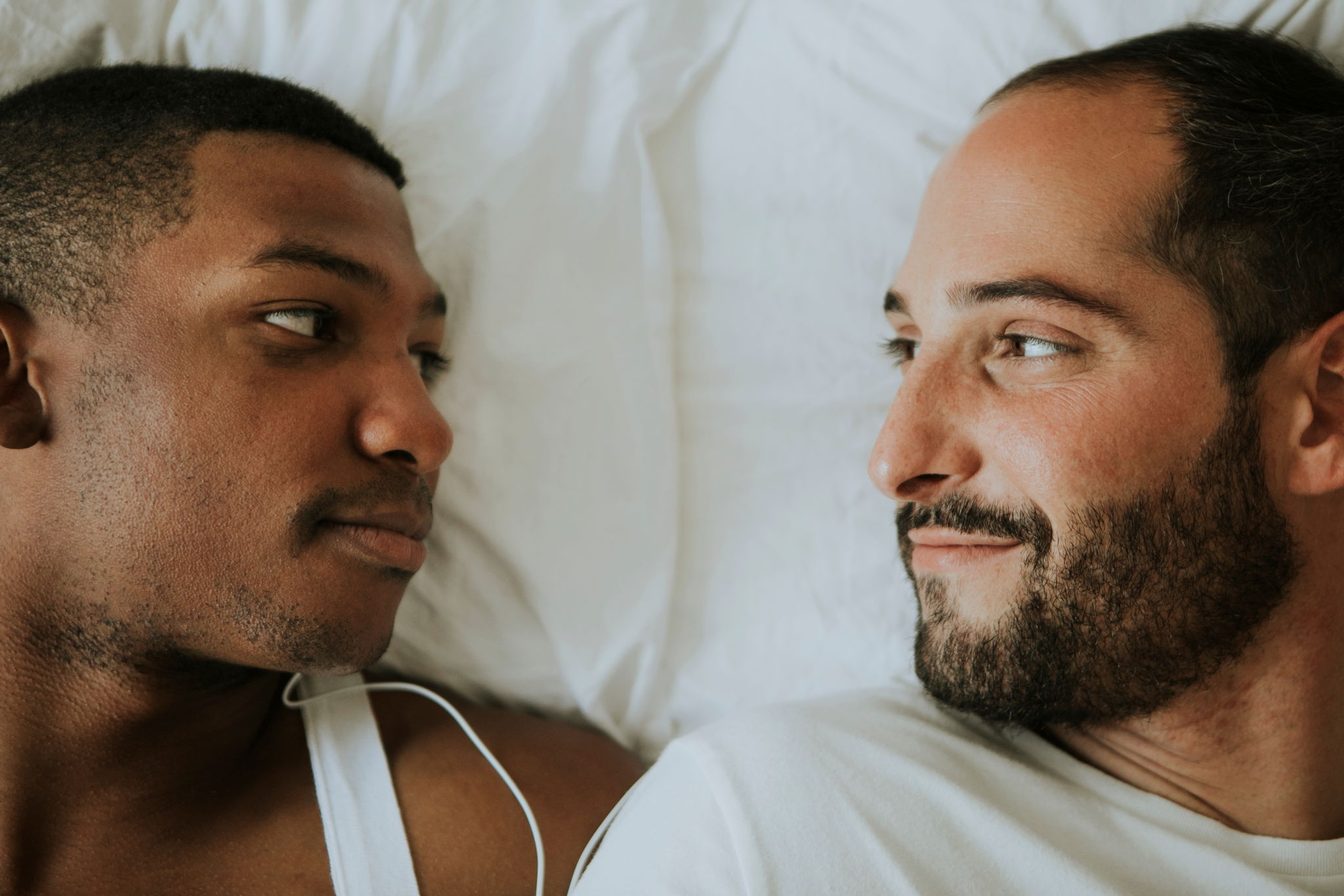 An attractive multi ethnic male couple lays in bed together smiling and looking at eachother.