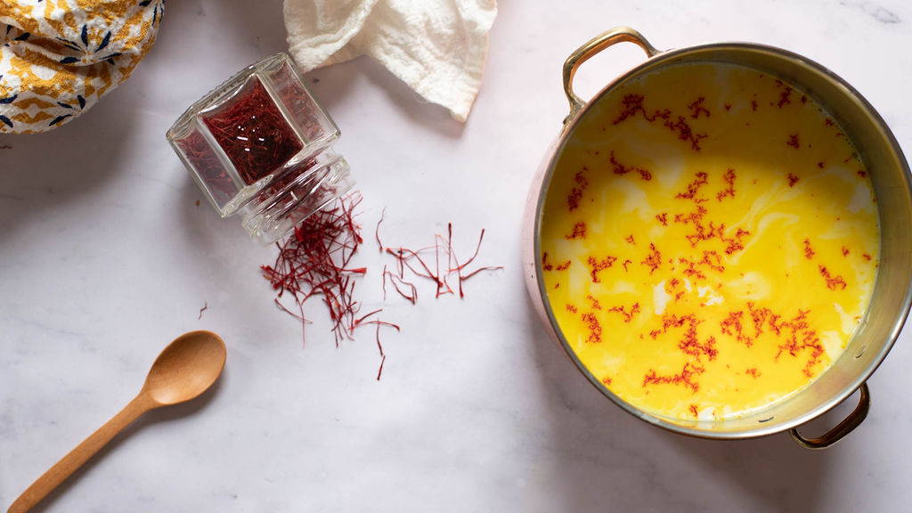 A glass jar of saffron is spilling saffron next to a metal pot full of a yellow liquid with saffron on top