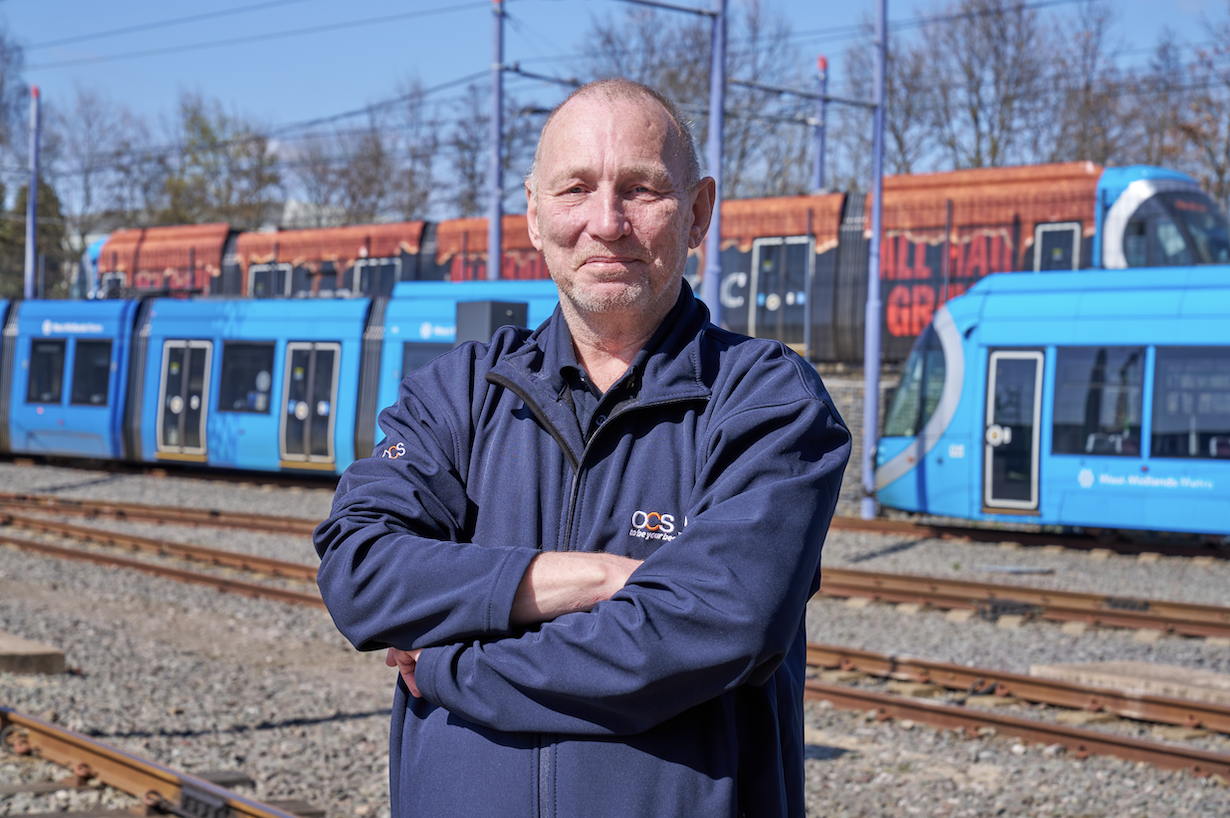 A man in a navy jacket stands with arms crossed on a railway track, with blue trams and a rust-colored train visible in the background on a sunny day.
