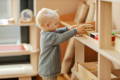 Little boy reaching towards his playroom shelf with wooden Montessori toys. 