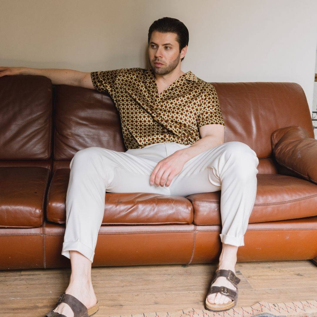 model sitting on a sofa wearing brown sandals white pants and a short sleeve gold bowling silk shirt from 1000 kingdoms