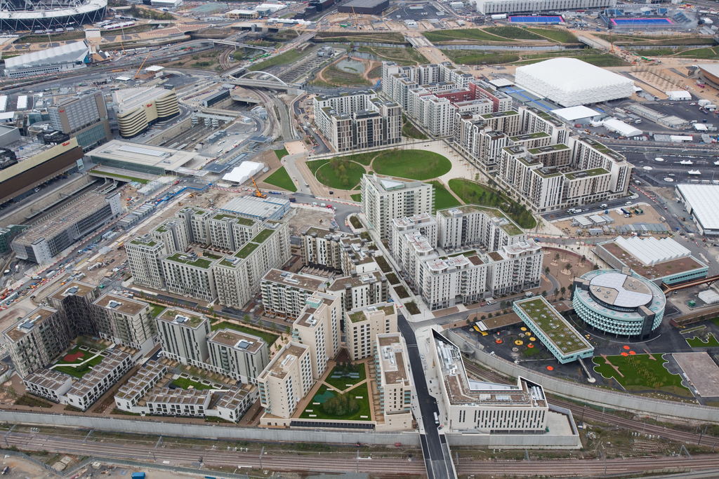 Aerial view of a modern urban development with multiple apartment buildings, green spaces, pathways, and nearby roads; surrounding areas are under construction and include stadiums and larger structures.