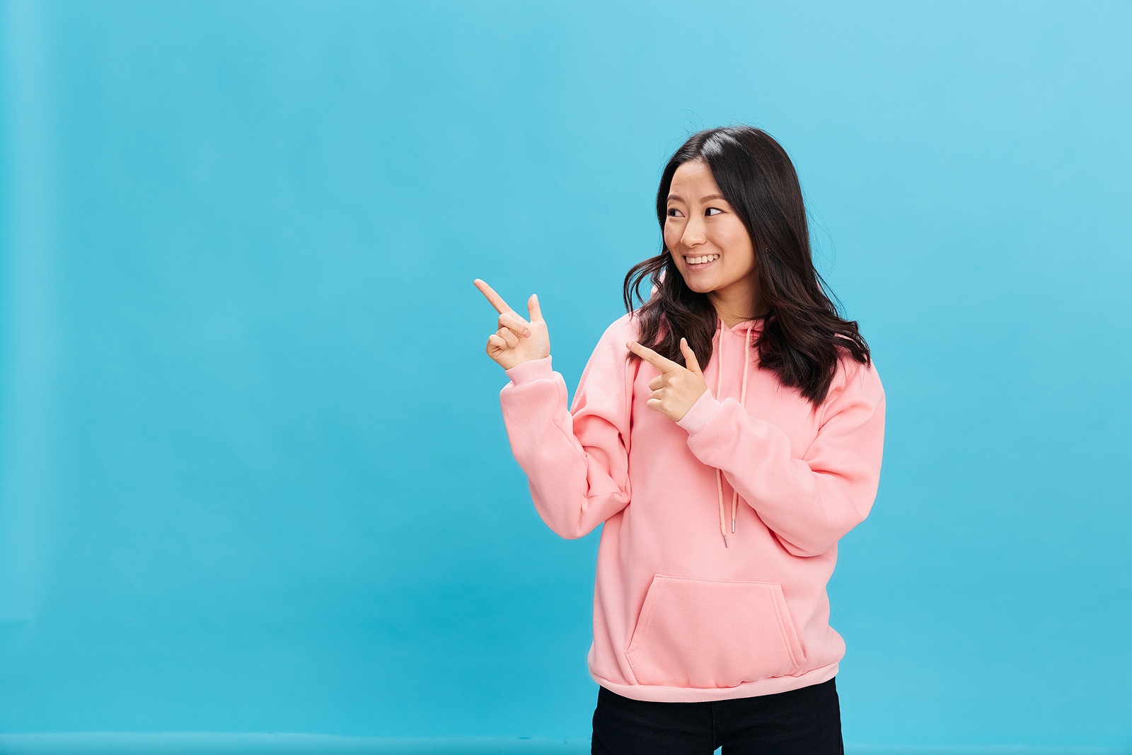 Overjoyed cute Asian student young lady in pink hoodie sweatshirt point finger to someone out of frame.