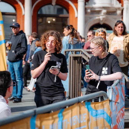 Two people in black t-shirts stand outside at an event, holding phones and speaking. They are surrounded by a crowd, with a building featuring arches in the background. It looks sunny and lively.