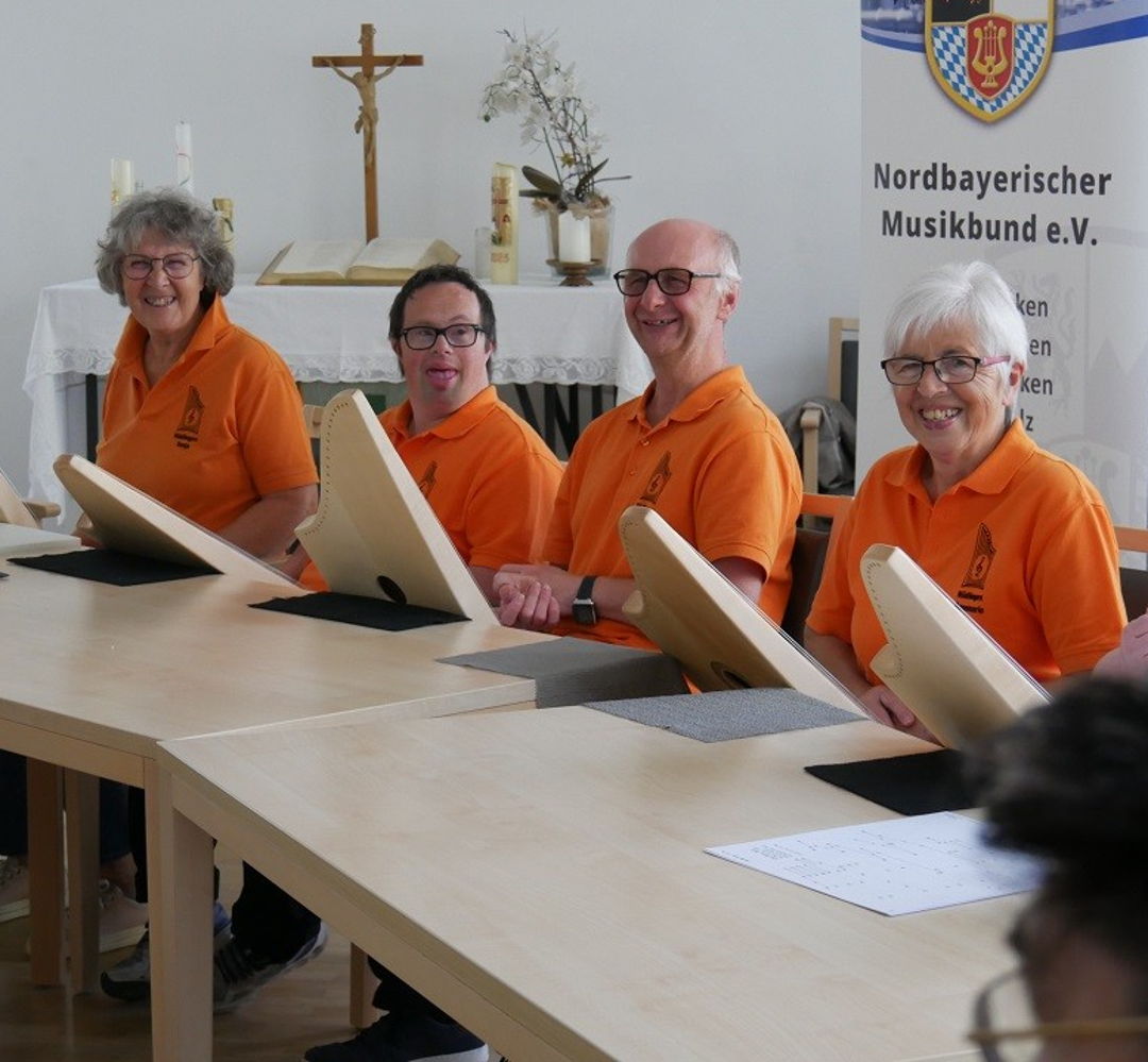 Fünf Menschen in orangenen T-Shirts sitzen an einem Tisch und spielen gemeinsam auf Tischharfen. 