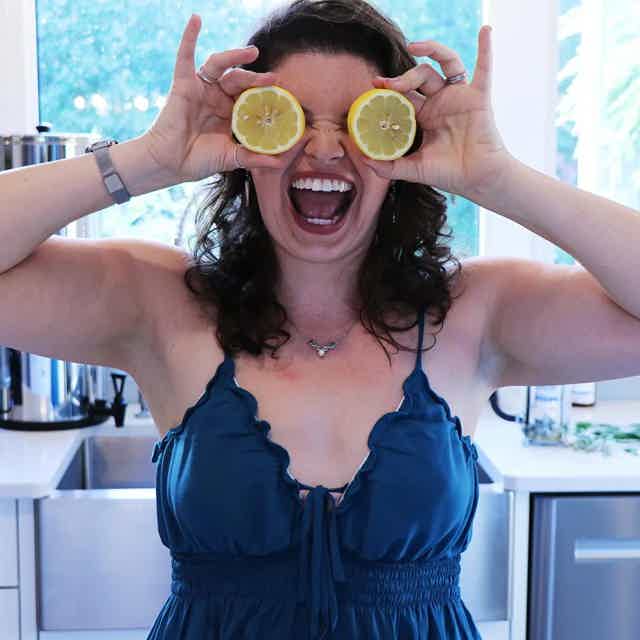 Woman in kitchen playfully holding lemon halves over her eyes, smiling widely, wearing a dark sleeveless top.