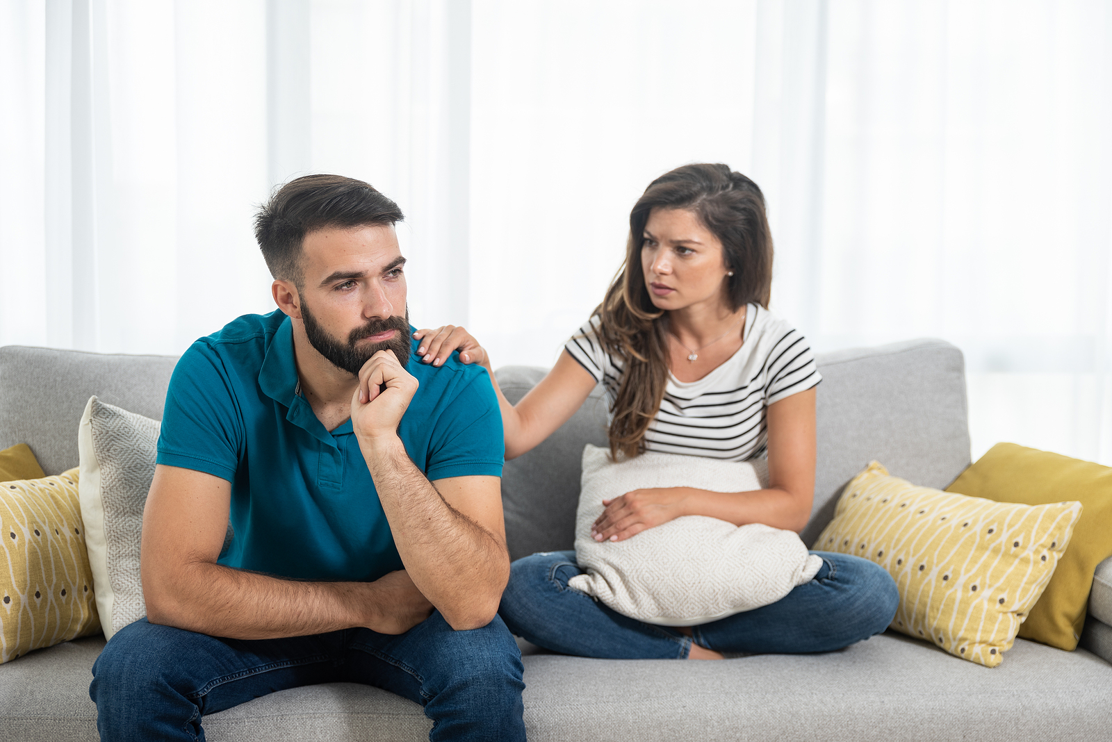 A young couple sits together on a couch, the man is thinking with his hand on his chin and the she is worried with her hand on him.