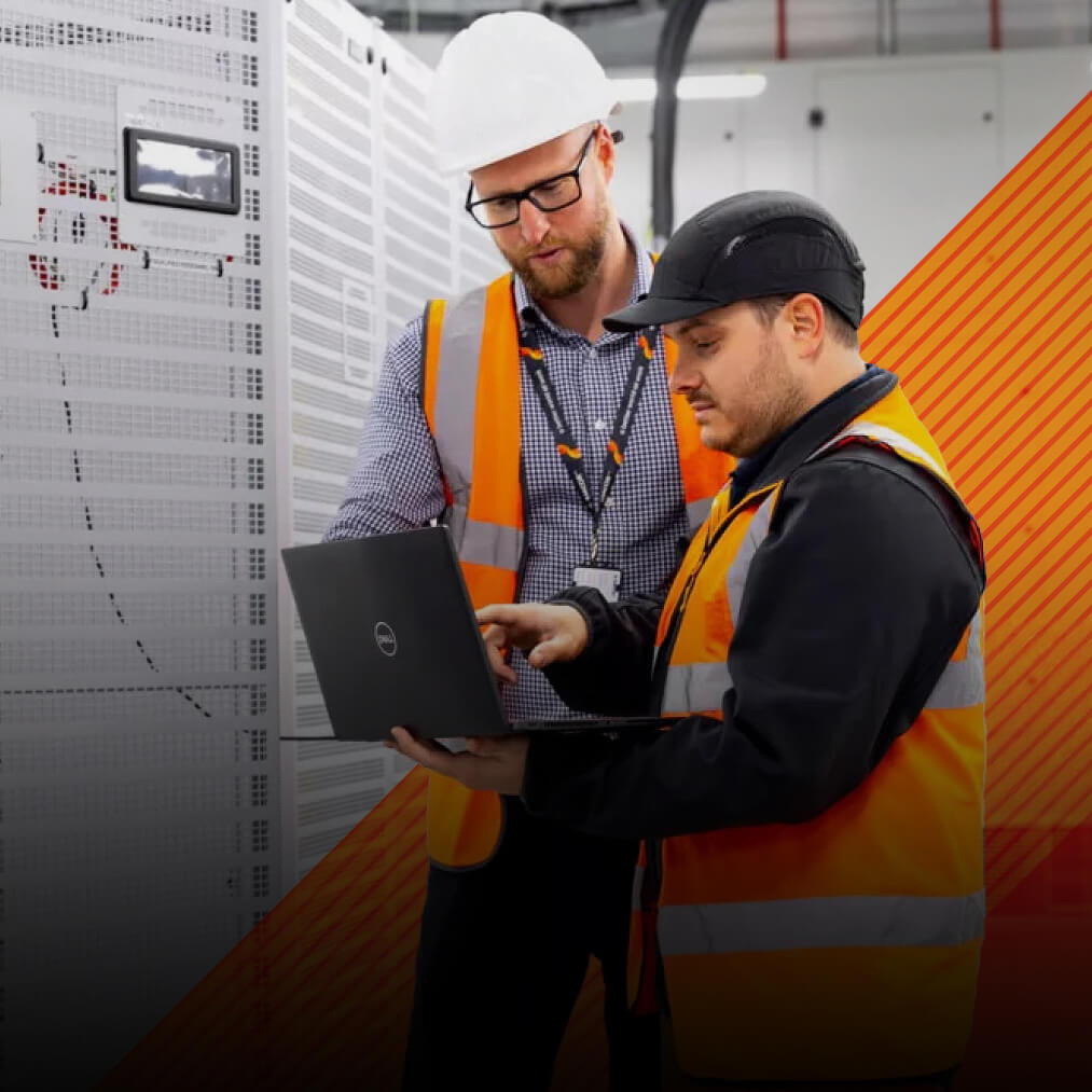 Two men wearing safety vests and hard hats work together in a server room. One holds a laptop while the other points at the screen. Tall racks of equipment are visible in the background.