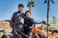 Ein älteres Paar steht lächelnd und mit Helmen auf hinter einem schwarzen und einem orangenen E-Bike. Im Hintergrund sind Palmen, Steine und blauer Himmel zu sehen.