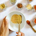 A person pours a yellowish ground spice into a white ceramic bowl which is surrounded by many open glass spice jars