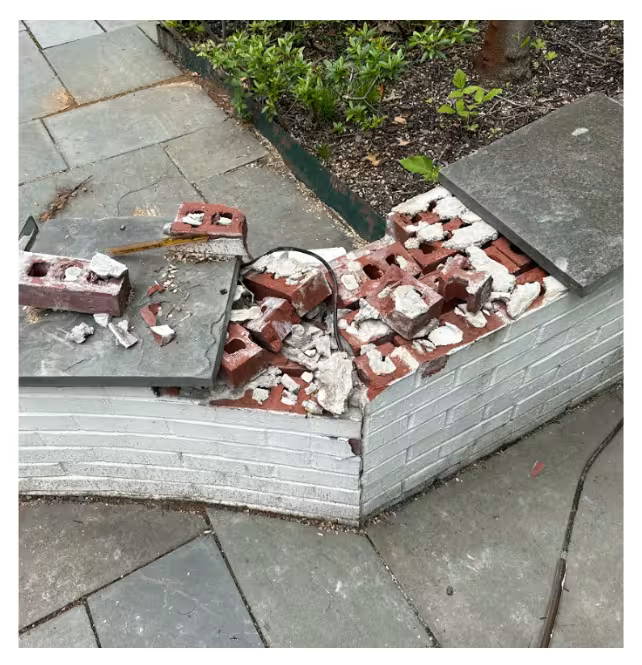 Damaged brick fire pit wall with collapsed masonry and broken bricks scattered on top, next to a stone patio and garden bed.