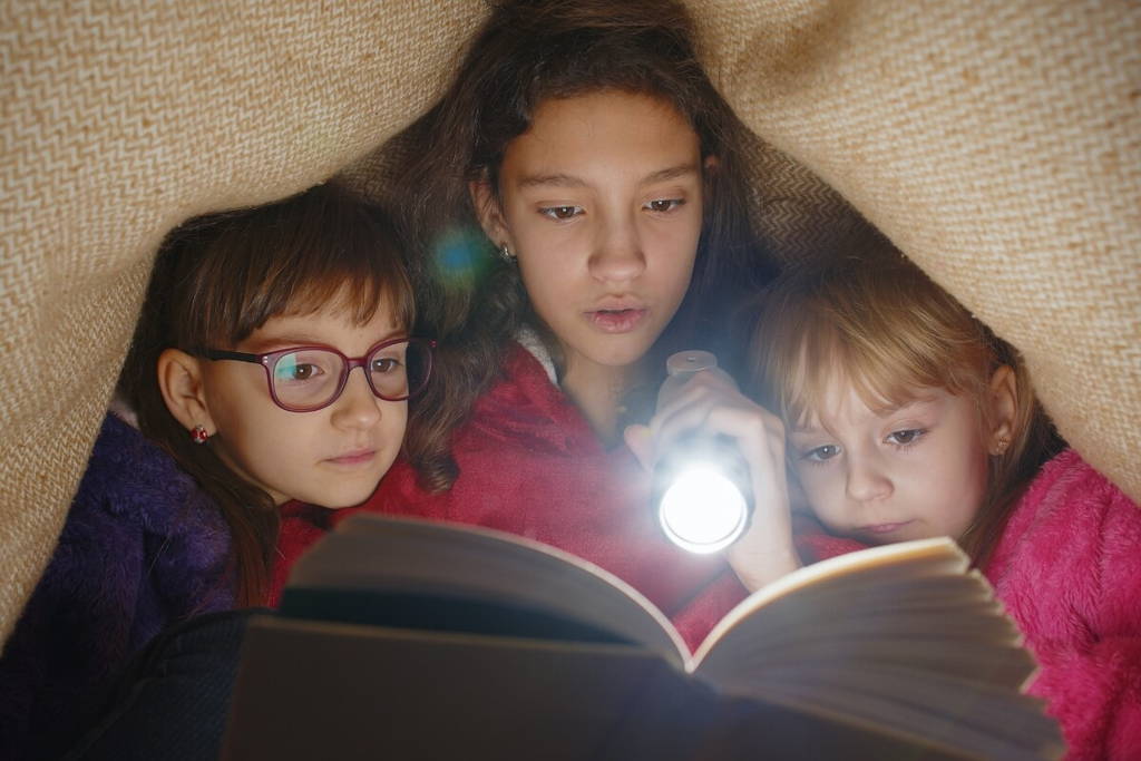 Three young girls reading a book together under a cozy blanket fort, with one girl holding a flashlight to illuminate the pages in a dim, intimate setting.
