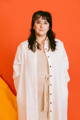 a woman stands with a soft smile and shoulder length brown hair with bangs in front of an orange background. She is wearing a full length white button up linen dress over a cream knit fitted shift. Her hands are in her pockets.