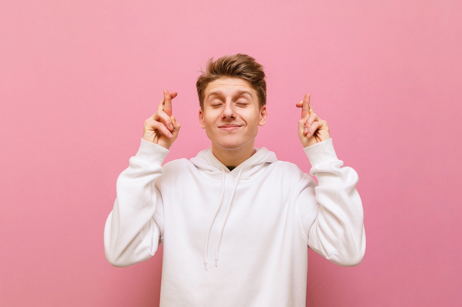 Positive young man thinking desire with crossed fingers and closed eyes against pink background.