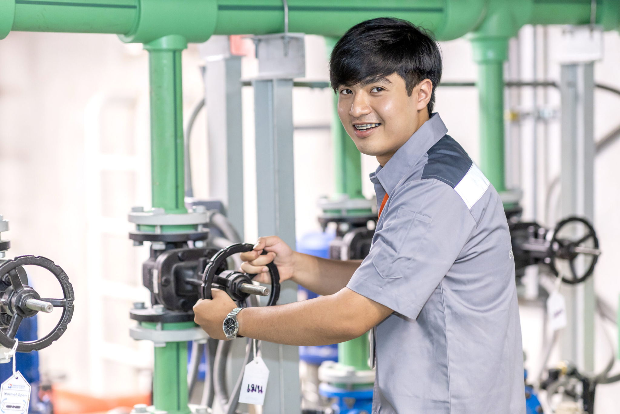 A young technician in a gray uniform smiles while operating a valve on industrial piping in a facility. Green and black pipes are visible in the background.