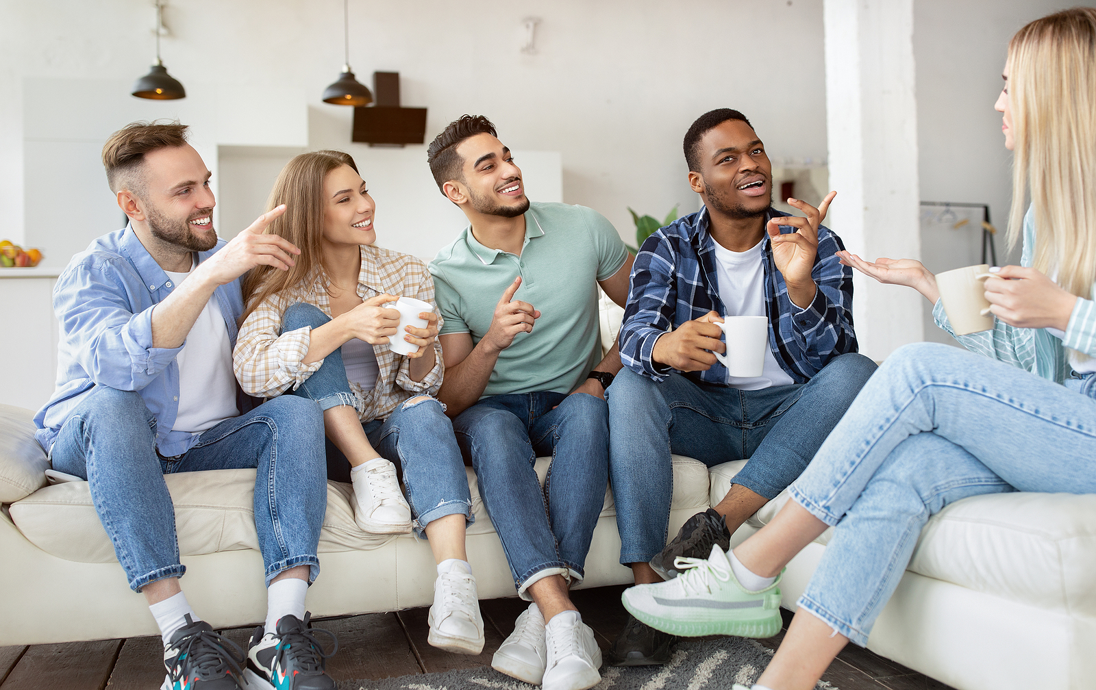 Image of several friends of different ethnicities sitting on a couch together with their coffee mugs asking questions to the blond haired woman on the right.