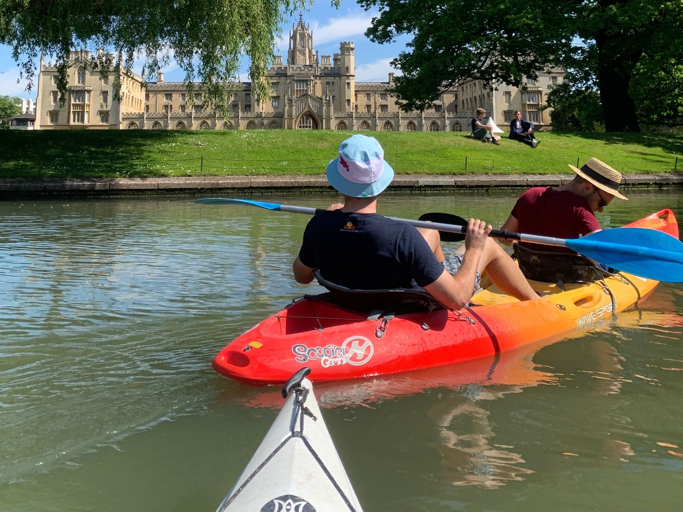 Cambridge university students kayaking on the river Cam overlooking a beautiful college building behind a patch of very green grass on the river bank. There are also two students sitting in the grass on the bank.