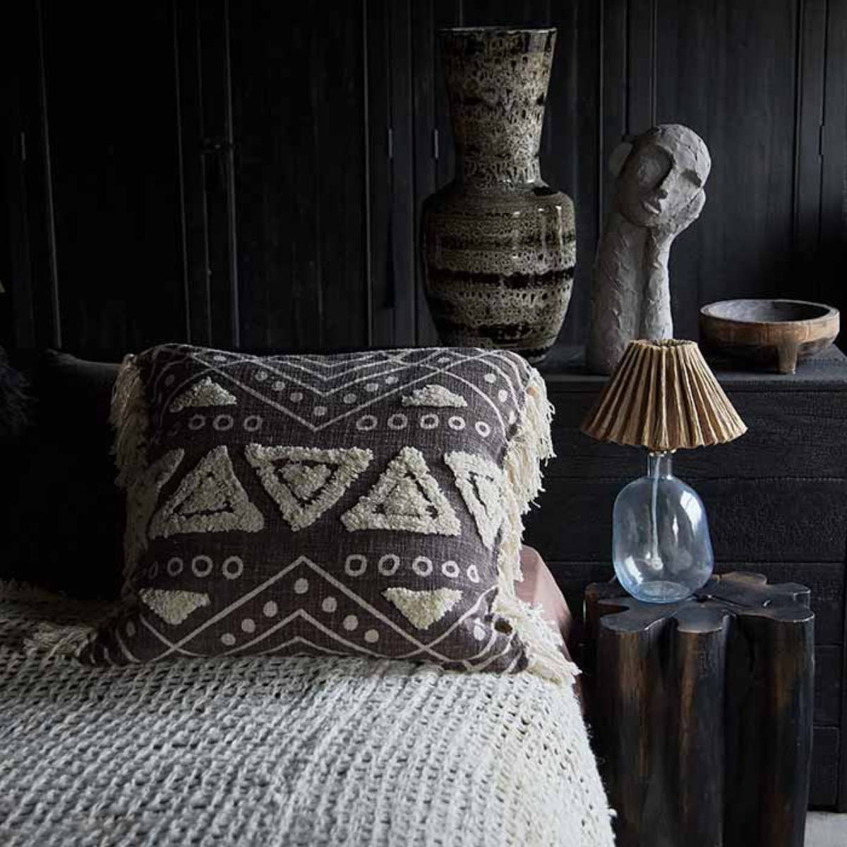 a bedside table and a large headboard housing a large ceramic German style vase next to a large head sculpture with a glass lamp with a pleated shade and a textured cushion that is brown with a raised cream pattern 